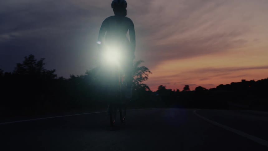 A silhouetted cyclist with a bright headlight rides down a road at sunset in slow motion