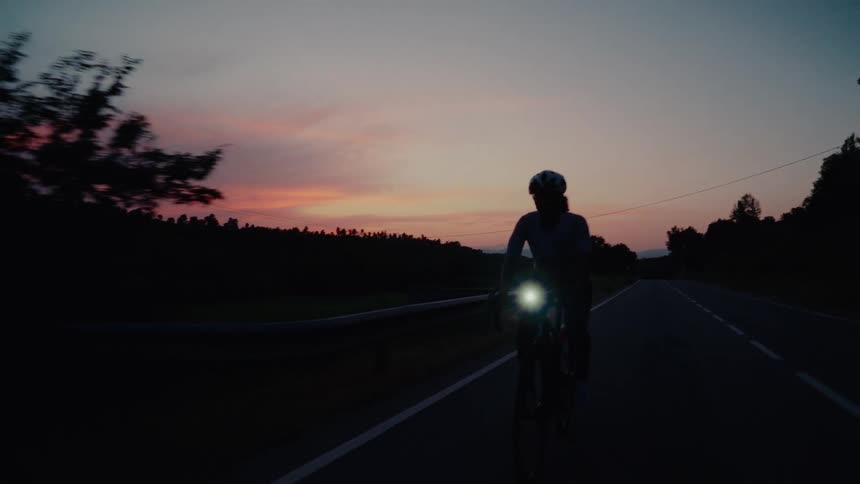 Front view of a determined cyclist pedaling on a rural road in the dark, illuminated by a headlight against a colorful dusk sky
