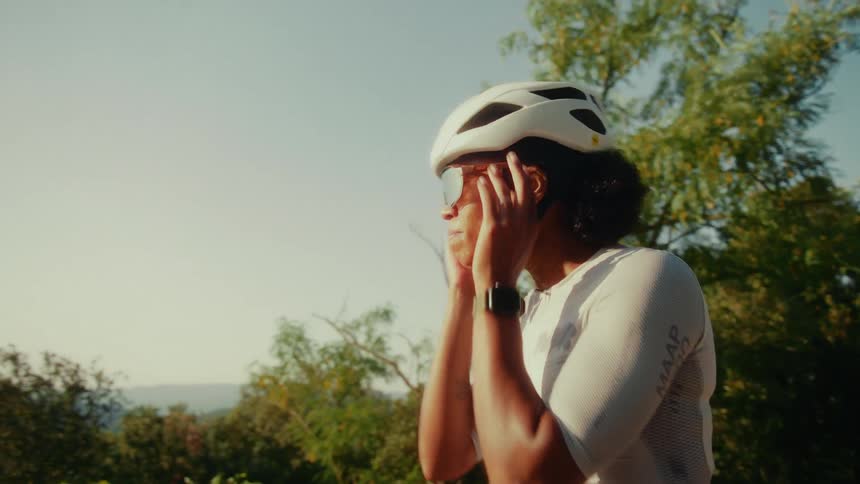 A smiling female cyclist in a white helmet puts on her sunglasses in slow motion on a beautiful sunny day