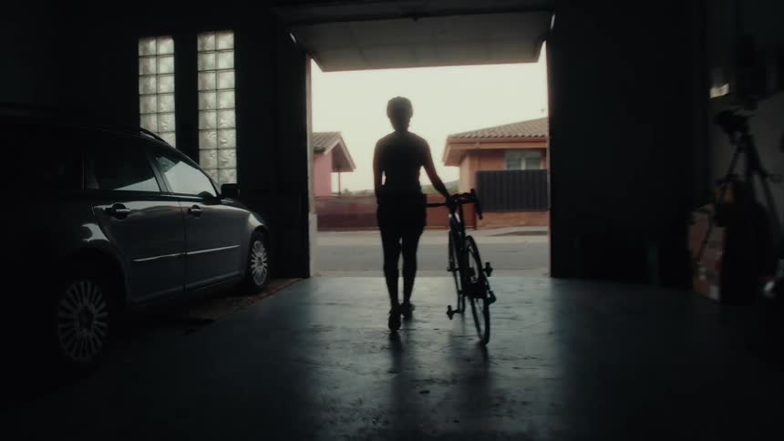 A determined female cyclist walks her bicycle out of a shadowy garage, silhouetted against the bright light outside