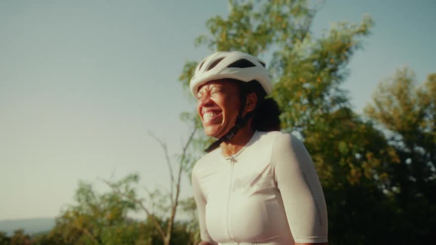 A joyful female cyclist in a helmet laughs heartily and wipes her brow in the warm sunshine
