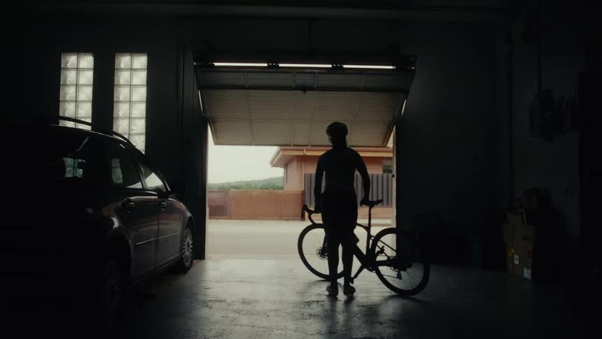 A silhouette of a cyclist in a helmet takes their bike and walks out of a dark garage to start their ride