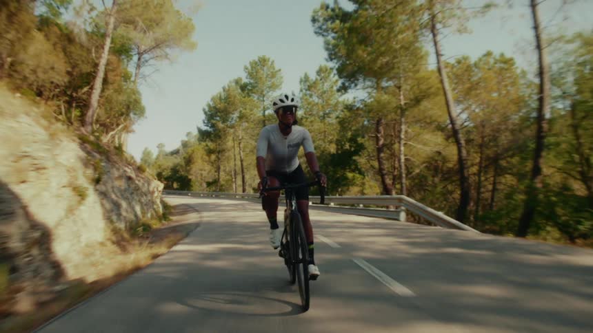 A determined female road cyclist in full gear rides her bike along a scenic, sunlit mountain road