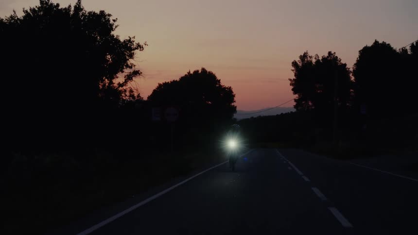 A cyclist with a bright headlight rides towards the camera on a dark country road at dusk