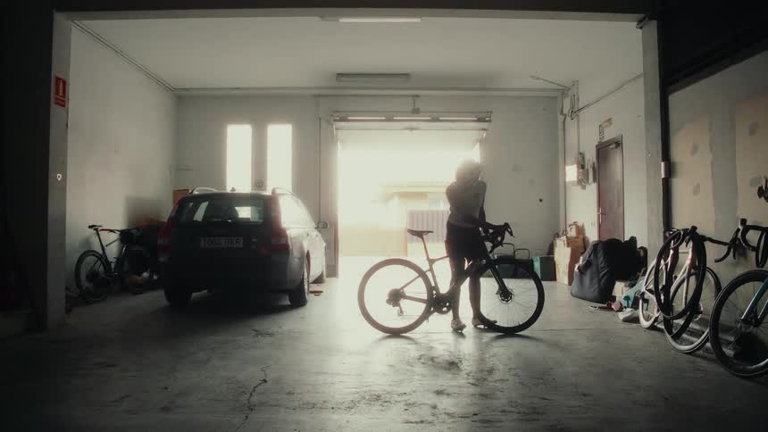 A female athlete in full cycling gear prepares her road bike inside a dark garage, silhouetted by the bright morning sun