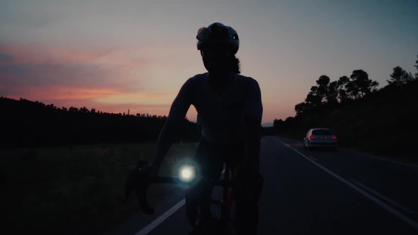 Slow-motion close-up of a professional cyclist riding on a dark road during a beautiful sunset