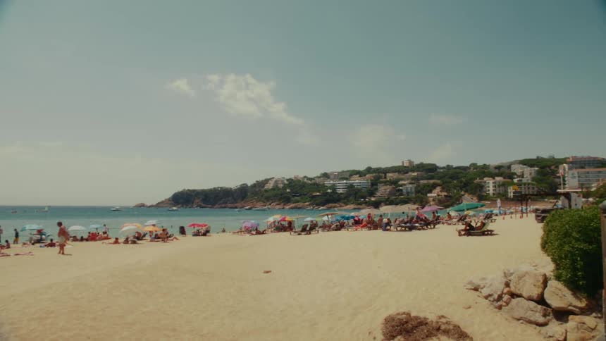 A static wide shot with a vintage feel shows a bustling European beach filled with sunbathers under colorful umbrellas