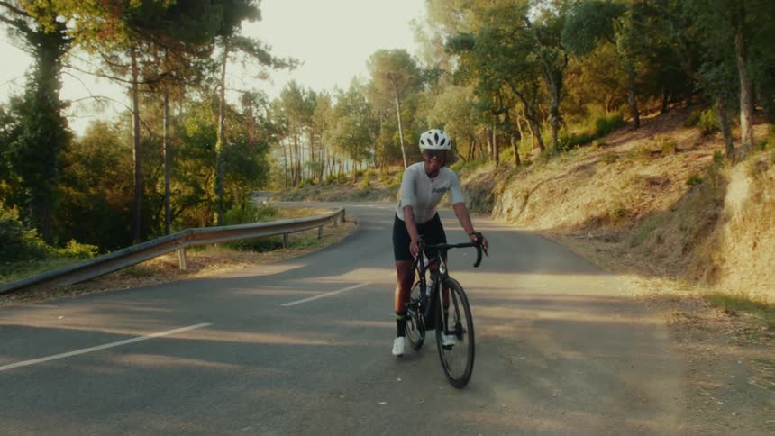 Slow motion of a happy female cyclist riding up a winding mountain road, surrounded by trees at golden hour