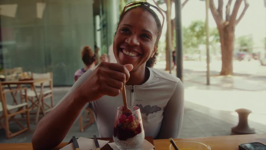 A joyful woman dressed in cycling gear smiles at the camera while stirring a vibrant fruit and granola parfait at a sunny outdoor cafe