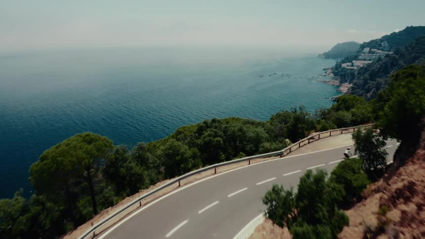Aerial drone shot follows a lone rider on an electric skateboard down a winding coastal road overlooking the vast blue ocean