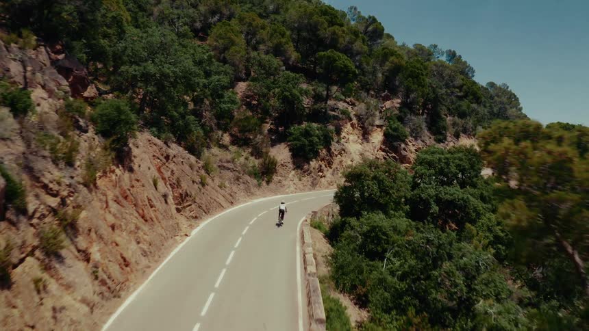 Aerial view of a cyclist pedaling up a winding road carved into a mountainside with dense green foliage