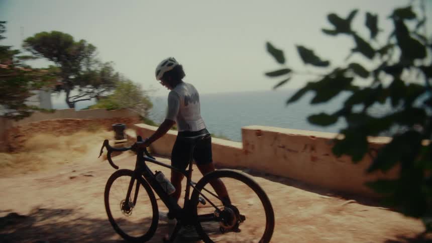 A female cyclist in full gear walks her bike along a scenic cliffside path with a beautiful ocean view