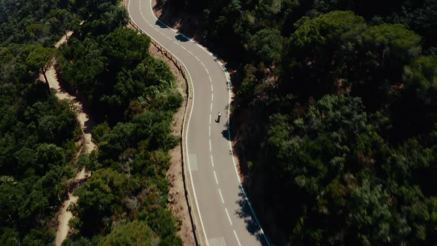 An aerial view captures a lone cyclist pedaling along a scenic, winding road that cuts through a dense, sunlit forest