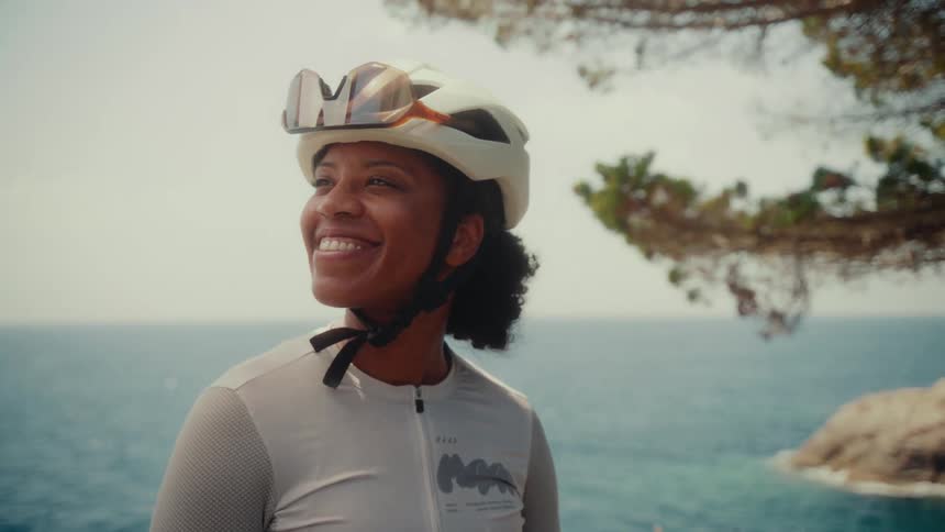 A happy female cyclist in professional gear smiles while enjoying a stunning ocean view from a clifftop