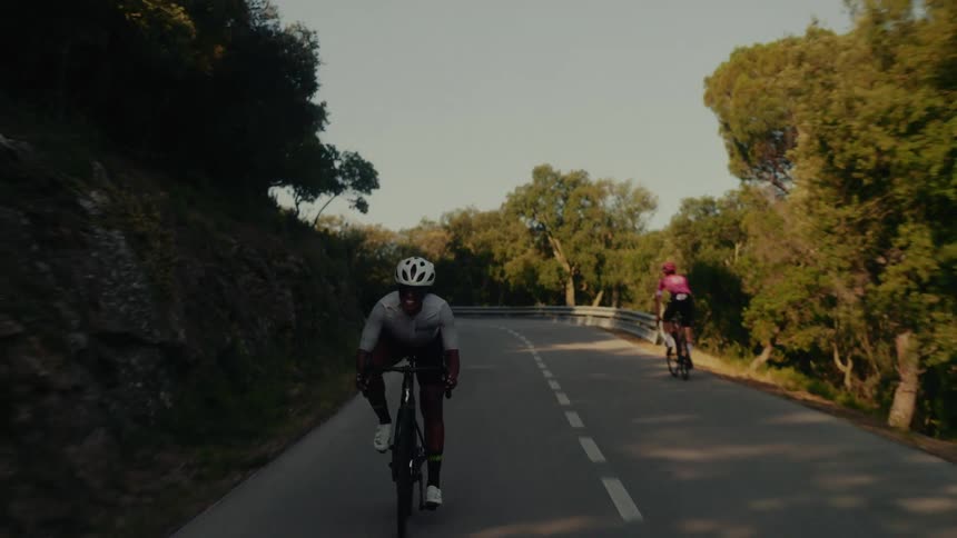 An athletic Black man grits his teeth in determination while cycling uphill on a winding road through a sun-drenched forest