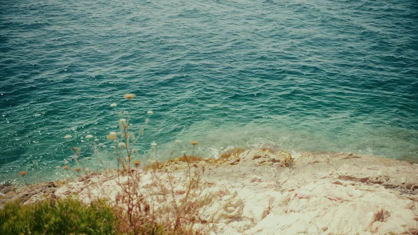 A serene, high-angle view of the turquoise ocean gently lapping against a rocky, sunlit shore on a warm summer day