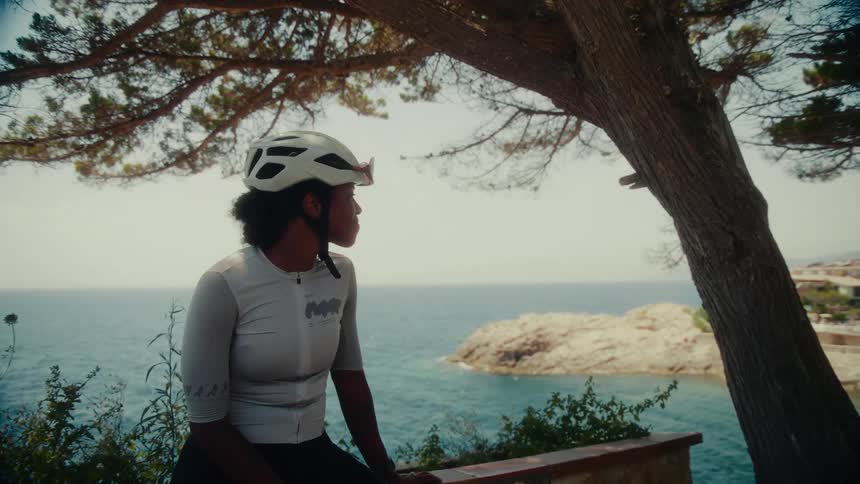 A female cyclist in a helmet rests under a tree and peacefully gazes at the ocean view