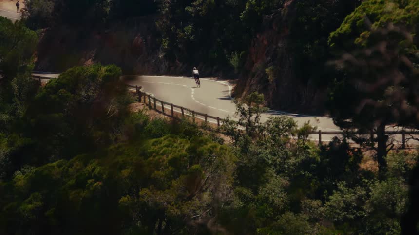 A lone cyclist rides up a winding road carved into a lush, green mountainside on a sunny day
