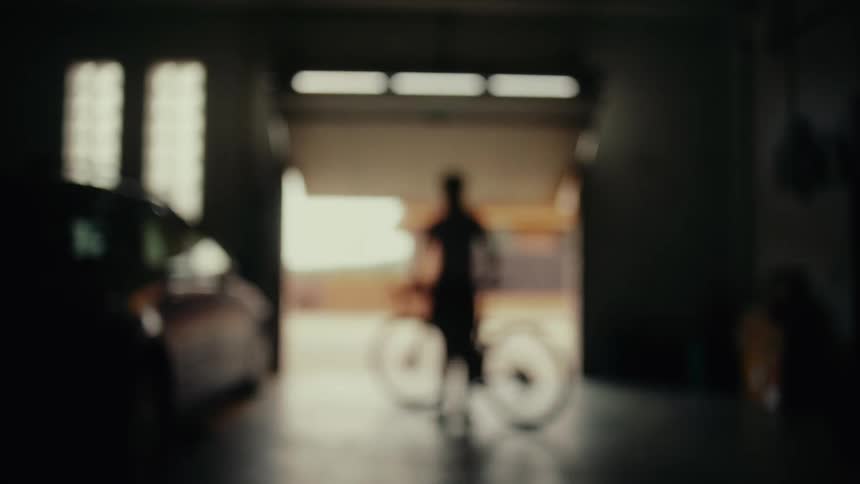 A blurry, silhouetted cyclist walks their bike out of a dark garage as the automatic door opens to the bright outdoors