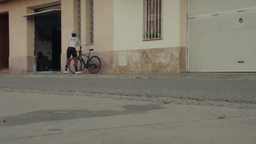 A female cyclist in full gear puts on her helmet and then walks her bicycle back into a building