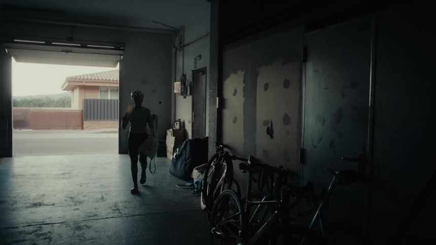 A silhouetted cyclist in full gear warms up by jogging in place inside a dark, open garage