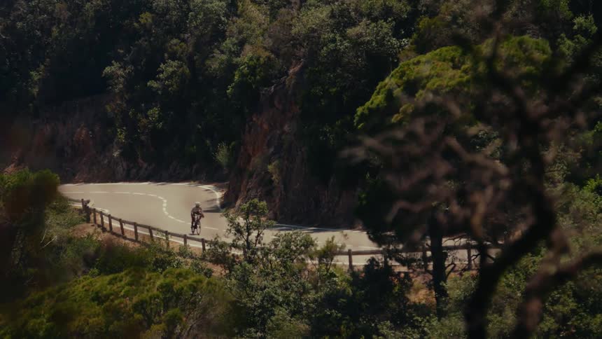High angle view of a cyclist navigating a winding mountain road through a dense, sun-drenched forest