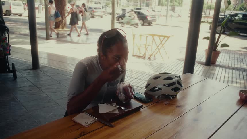 A female cyclist in athletic wear enjoys a healthy snack at a sunlit cafe with her helmet on the table