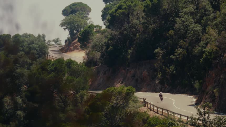 A lone cyclist descends a winding mountain road through a lush green forest on a foggy day