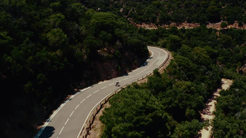Aerial footage follows a lone cyclist navigating a winding paved road through a dense, sun-drenched green forest
