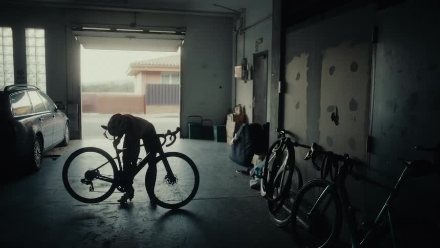 A silhouetted female cyclist in full gear prepares her road bike inside a dark garage before heading out for a ride
