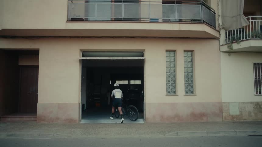 A cyclist wearing a helmet and full gear wheels their road bike into an open residential garage