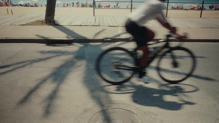 A cyclist rides through the prominent shadow of a palm tree on a sunny boardwalk next to a busy beach
