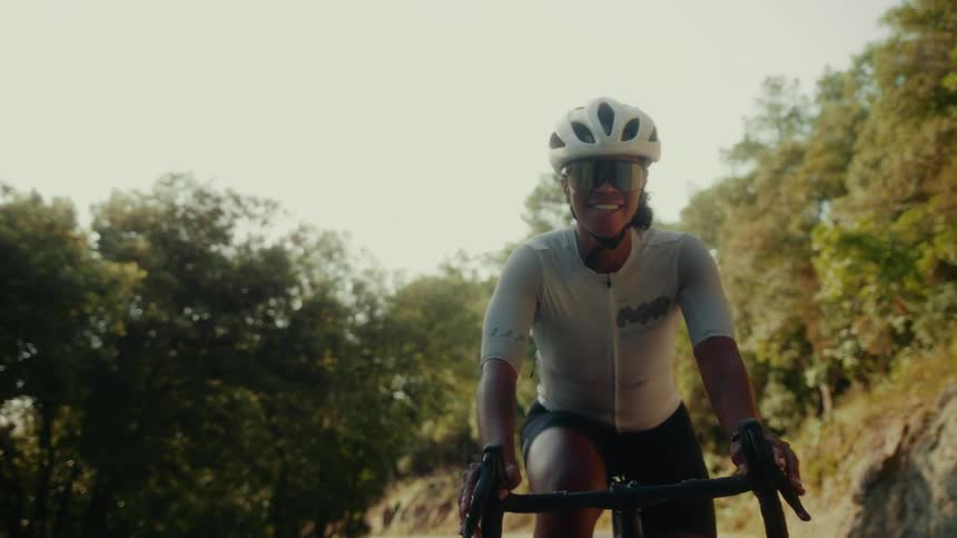 A smiling female cyclist in full gear rides her road bike uphill through a sun-drenched forest in slow motion