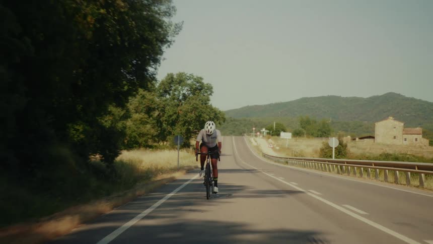 An athletic cyclist rides a road bike down an empty highway through the sunny countryside