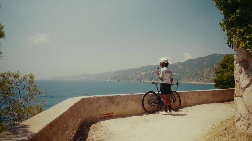 A professional cyclist takes a moment to rehydrate and enjoy the stunning Mediterranean sea view from a scenic overlook