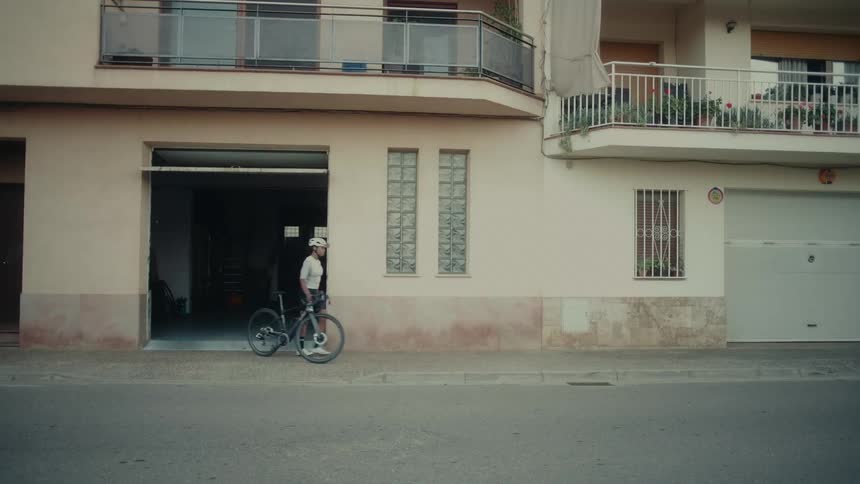 A woman in cycling gear walks her bike out of a garage and onto the sidewalk to start her ride
