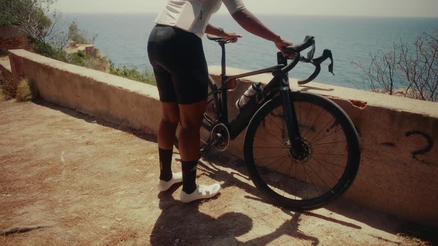 A cyclist stands with their bike overlooking the calm sea on a sunny day in a cinemagraph