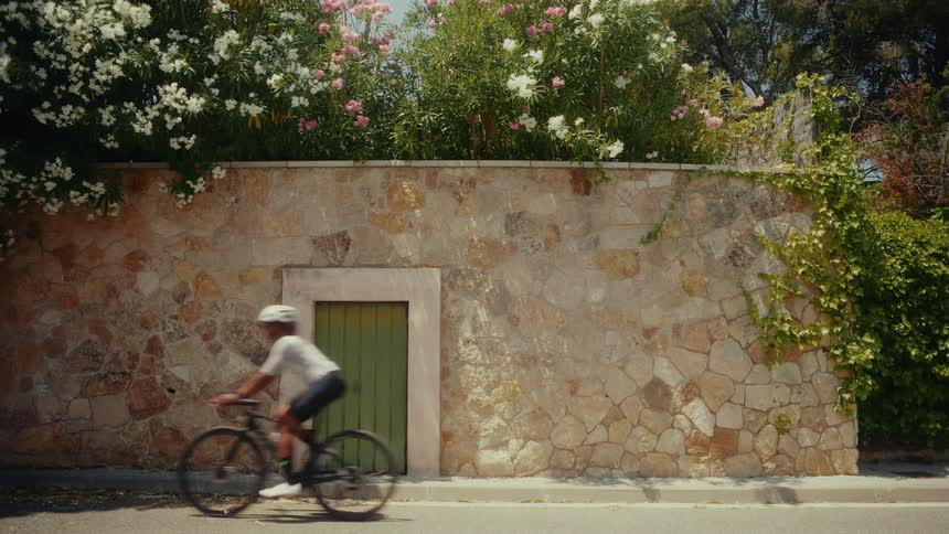 A cyclist rides past a beautiful stone wall topped with blooming flowers on a sunny day