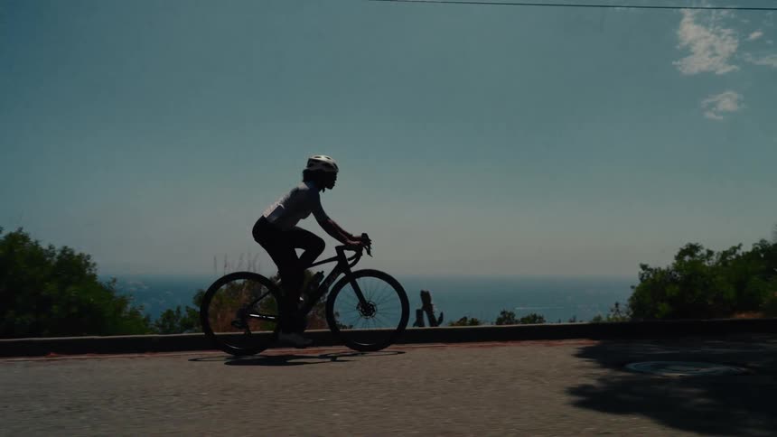 A silhouetted female cyclist in slow motion rides her road bike up a coastal mountain road with a beautiful ocean view