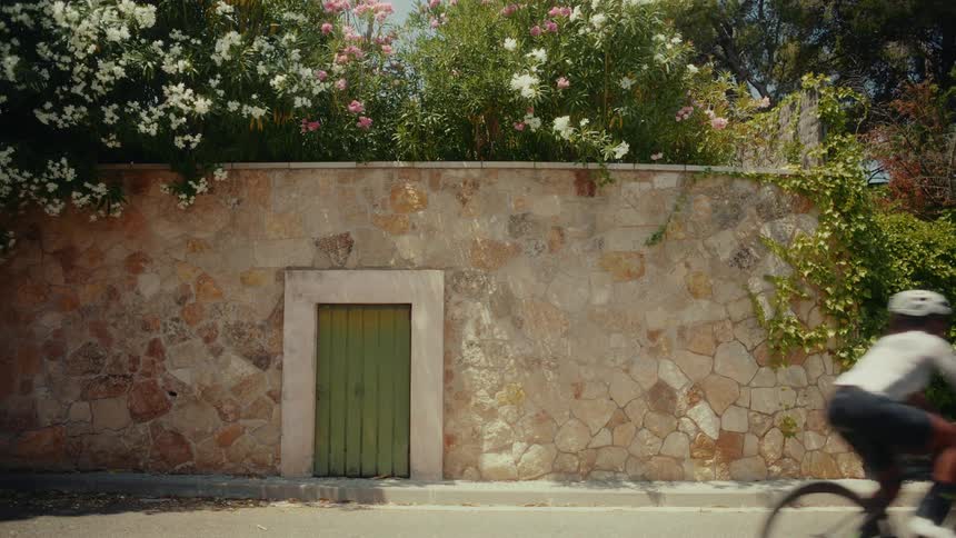 A cyclist rides swiftly past a picturesque stone wall adorned with blooming flowers on a sunny day