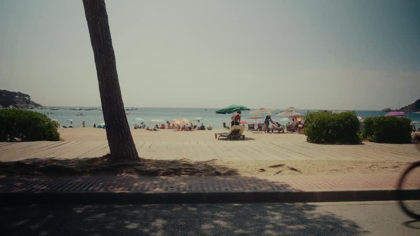 A blurry cyclist speeds past a sunny beach scene with people relaxing under umbrellas by the ocean