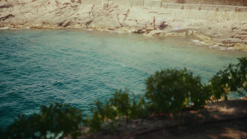 A tranquil high-angle view shows people relaxing on a secluded rocky cove with clear turquoise water on a sunny day
