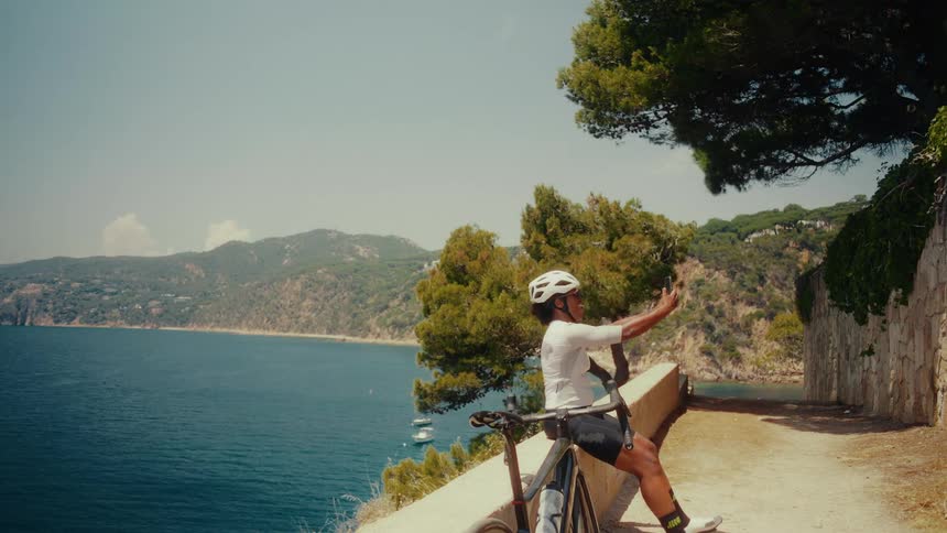 A female cyclist takes a break on a scenic coastal ride to sit down and capture a selfie with the stunning ocean backdrop