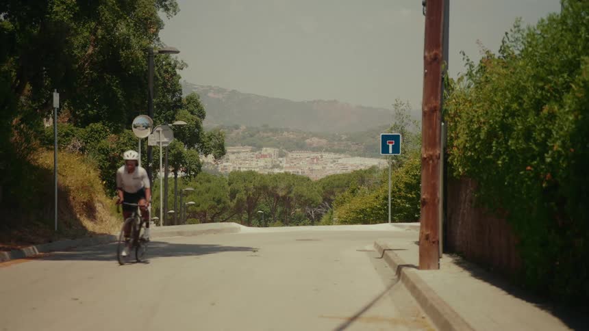 A woman of color wearing a helmet and cycling gear rides her road bike uphill on a scenic road with a town in the background