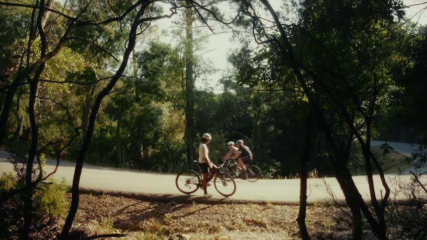 A cyclist takes a break on a sun-drenched, tree-lined road while other cyclists ride past in the background