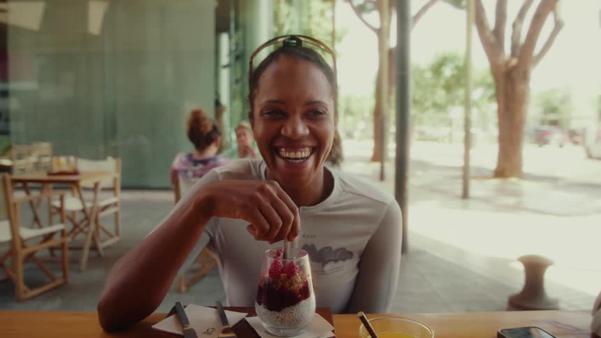 A joyful female cyclist laughs while enjoying a healthy parfait at a sunny outdoor cafe