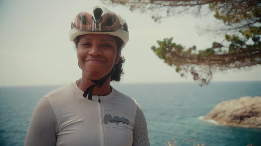 An empowered female cyclist in full gear smiles warmly at the camera with a beautiful ocean view in the background