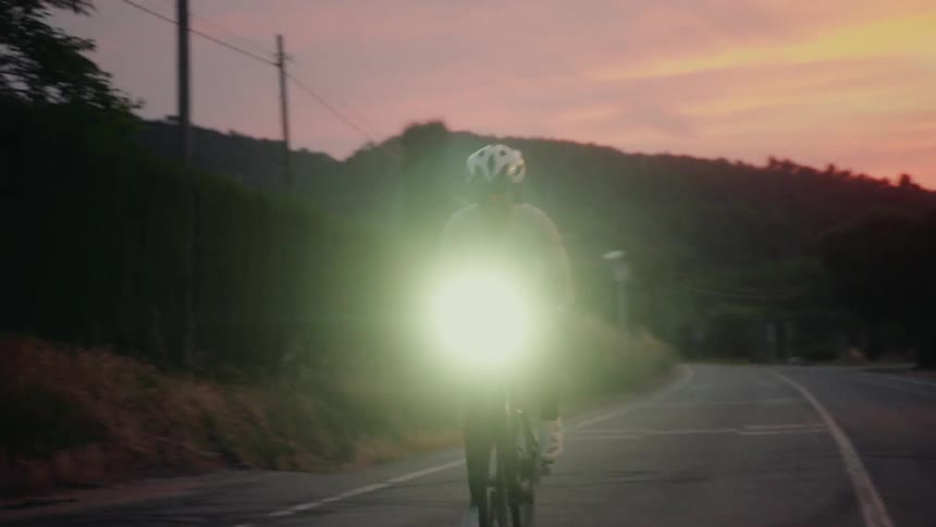 A joyful cyclist rides their bike in slow motion towards the camera on a rural road during a colorful sunset, their bright headlamp lighting the way