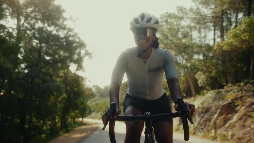 A smiling female cyclist in professional gear rides her road bike down a sun-drenched country road in slow motion