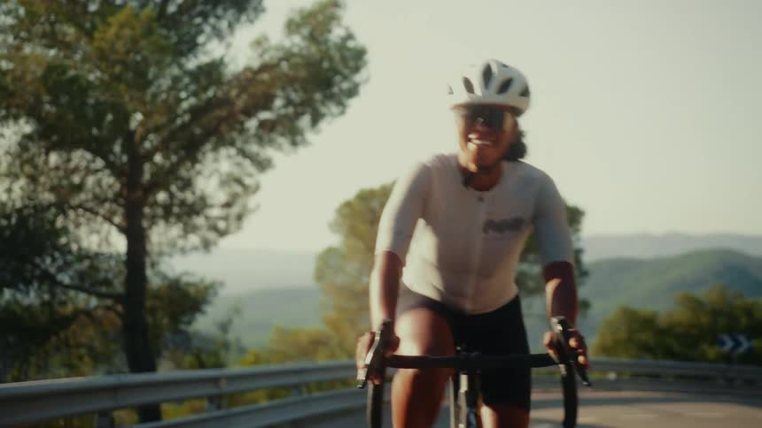 In slow motion, a professional female cyclist smiles as she rides her road bike up a scenic, winding mountain road on a sunny day
